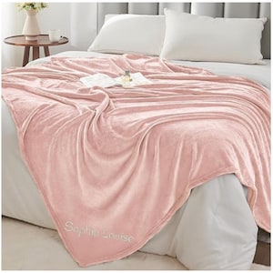 May include: A soft, blush pink blanket draped over a bed. The blanket has a velvety texture and is embroidered with the name "Sophia Louise" in a delicate script. A book and perfume bottle rest on top of the blanket.