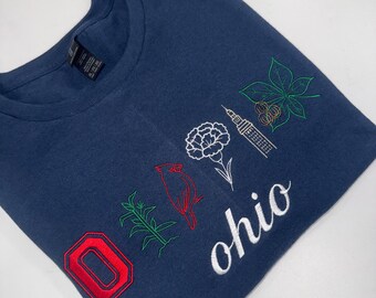 May include: Navy blue t-shirt with embroidered Ohio state symbols. Features a red "O" patch, a cardinal, a white carnation, a building, a green leaf, and the word "ohio" in white script.