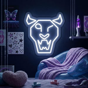 May include: A white neon sign shaped like a winking lion's head is fixed to a dark wall. A clear plastic chair with a purple blanket is positioned below the sign. Other items include a lava lamp and artwork. The sign emits a bright glow.