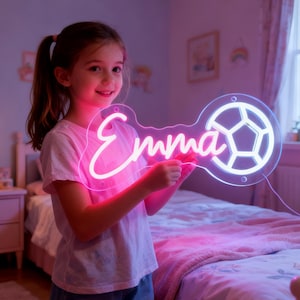May include: A pink neon sign in the shape of a key, featuring the name "Emma" in cursive and a soccer ball design. The sign is held by a person in a pink shirt, with a bedroom setting in the background.