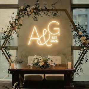 May include: A wedding reception table is decorated with floral arrangements, candles, and a neon sign that reads "A&G EST 2026". The table is made of dark wood and has two benches. The background features a geometric wooden arch adorned with flowers and greenery.