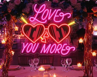 May include: A neon sign with the words "Love You More" in pink script. The sign features heart-shaped sunglasses with an arrow through them. The sign is part of a wedding or event decoration.