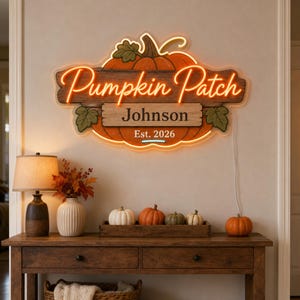 Op de afbeelding: Een aan de muur gemonteerd neonbord in de vorm van een pompoen met oranje oplichtende contouren. Het bord bevat de tekst Pumpkin Patch in fel oranje neon, met de gedrukte tekst Johnson en Est. 2026 op een houtlook plank met groene bladeren.