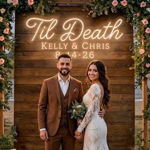 Peut inclure: Un couple pose devant un fond en bois orné de compositions florales et d'une enseigne au néon qui dit "Til Death". L'enseigne affiche également les noms "Kelly & Chris" et la date "8-14-26". L'homme porte un costume marron et la femme porte une robe de mariée en dentelle blanche.