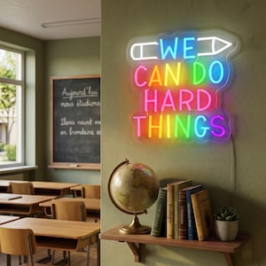 Peut inclure: Une scène de classe avec une enseigne au néon qui dit "WE CAN DO HARD THINGS" en couleurs arc-en-ciel. Un globe, des livres et une petite plante en pot sont sur une étagère en bois. Des bureaux et un tableau noir sont également visibles.