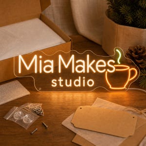 以下が含まれることがあります： 温かみのあるオレンジ色の光でMia Makes studioという文字が浮かび上がる透明なアクリル製LEDネオンサイン。緑色の湯気が立ち上るオレンジ色のコーヒーカップのアイコンが添えられています。取り付け金具と段ボール箱が付属しています。