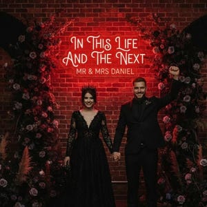 May include: A newly married couple pose in front of a brick wall with a neon sign that reads "In This Life And The Next" and "MR & MRS DANIEL". The woman wears a black gown and crown, and the man wears a black suit.
