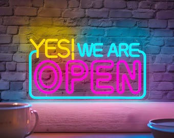 May include: A neon sign with the words "YES! WE ARE OPEN" in bright yellow, blue, and pink. The sign is rectangular and mounted on a brick wall. The sign is lit up, indicating the business is open.