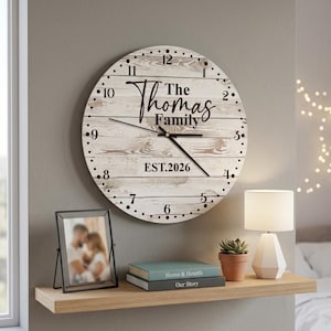 Könnte beinhalten: Eine runde Wanduhr mit weiß gekalkter Holzmaserung, schwarzen Ziffern und schwarzen Zeigern. In der Mitte steht der Text "The Thomas Family" und "EST.2026" in einer schwarzen Schreib- und serifenlosen Schrift. Die Uhr ist kreisförmig.