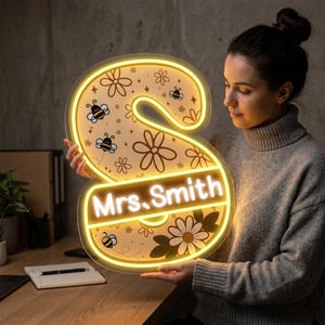 May include: A personalized illuminated sign in the shape of the letter "S". The sign features a floral and bee design with the name "Mrs. Smith" in white lettering. The sign has a warm yellow glow.