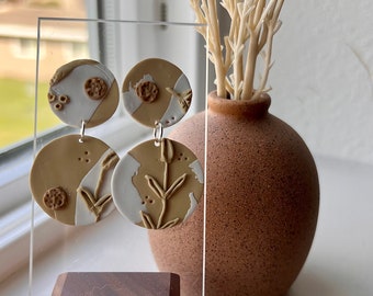 May include: A pair of handmade polymer clay earrings with a brown and white abstract design. The earrings are displayed on a wooden stand.
