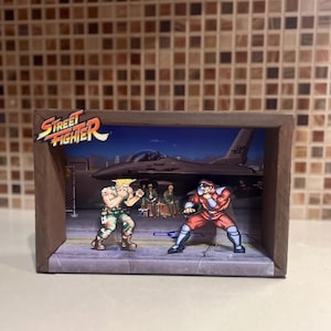 Puede incluir: Un diorama de una escena del videojuego Street Fighter. El diorama presenta dos personajes, Guile y Ryu, luchando en un escenario de base militar. El diorama está hecho de cartón e incluye un pequeño modelo de avión.