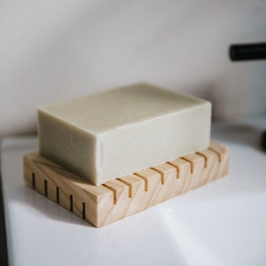 May include: A light green bar of soap resting on a wooden soap dish with grooves. The soap dish is made of light brown wood.