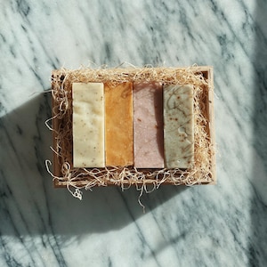 May include: Four bars of handmade soap in a wooden box with shredded paper. The soaps are in shades of white, yellow, pink, and green.