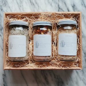 May include: Three glass jars filled with bath salts in a wooden box with shredded paper. The jars have white labels with the text "HONEYWILD BOTANICAL BATH SALTS" and the size of the product.