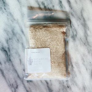 May include: A clear plastic bag filled with beige botanical bath salts. A white label with the text "HONEYWILD" and "BOTANICAL BATH SALTS" is attached to the bag. The bag is resting on a marble surface.