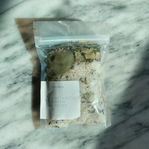 May include: A clear plastic bag filled with white bath salts and dried herbs. The bag has a white label with the text "HONEYWILD BOTANICAL BATH SALTS".
