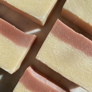 May include: Four bars of soap with a white base and a pink stripe on top. The soap bars are arranged in a cross pattern on a white marble surface.