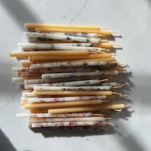 May include: A collection of thin, cylindrical taper candles stacked horizontally. The candles are a mix of solid yellow beeswax and white wax embedded with dried botanical pieces, including green herbs and small pink flower petals.