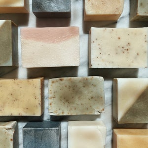 May include: Assortment of handmade soap bars in various colors and textures, including beige, brown, gray, and pink. The bars are arranged on a white marble surface.
