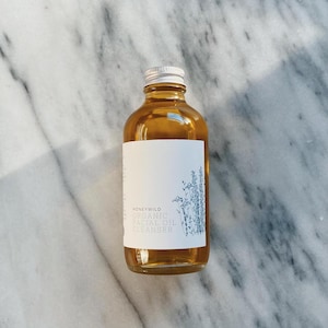 May include: A clear glass bottle with a silver lid filled with a golden liquid. The bottle has a white label with the text "HONEYWILD ORGANIC FACIAL OIL CLEANSER" and a small illustration of a plant.