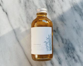 May include: A clear glass bottle with a silver lid filled with a golden liquid. The bottle has a white label with the text "HONEYWILD ORGANIC FACIAL OIL CLEANSER" and a small illustration of a plant.