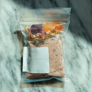 May include: A clear plastic bag filled with pink bath salts and dried flowers. The bag has a white label that reads "Honeywild Botanical Bath Salts, Net Weight 1.4 oz."