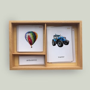 Könnte beinhalten: Eine Holzkiste mit drei Fächern, jedes mit einer Karte mit einem Bild und einem Wort. Das obere Fach hat eine Karte mit einem Bild eines Heißluftballons und dem Wort "Ambulanz". Das mittlere Fach hat eine Karte mit einem Bild eines Traktors und dem Wort "Traktor". Das untere Fach hat eine Karte mit einem Bild eines Heißluftballons und dem Wort "Ambulanz".