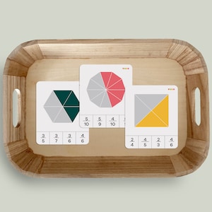 Pode incluir: Três cartões com diferentes formas geométricas coloridas divididas em frações. As frações são escritas abaixo de cada forma. Os cartões estão em uma bandeja de madeira.