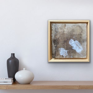 Op de afbeelding: Een ingelijst abstract schilderij met een lichtbruine achtergrond en witte verfspatten. Het schilderij is in een licht houten lijst en hangt aan een witte muur.