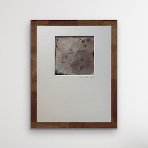Op de afbeelding: Abstract kunstwerk in een houten lijst. Het kunstwerk heeft een vierkante afbeelding met een getextureerde, gevlekte uitstraling in tinten bruin, beige en paars. De lijst is van donker hout met een zichtbare nerf. Het kunstwerk is voorzien van een witte rand.