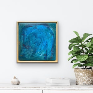 Puede incluir: Pintura abstracta en tonos azules y verdes, enmarcada en oro, colgada en una pared blanca.