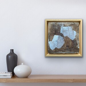 Op de afbeelding: Een ingelijst abstract schilderij met een gouden lijst. Het schilderij heeft twee witte vormen op een bruine achtergrond.