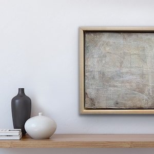May include: A framed abstract painting with a white background and subtle grey and brown brushstrokes. The painting is displayed on a wooden shelf with a black vase and a white ceramic vase.