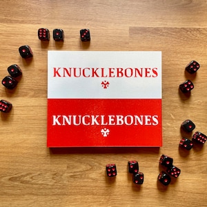 Może przedstawiać: Czerwono-białe pudełko z nadrukiem "KnuckleBones". Pudełko jest otoczone czarnymi kostkami do gry z czerwonymi kropkami.