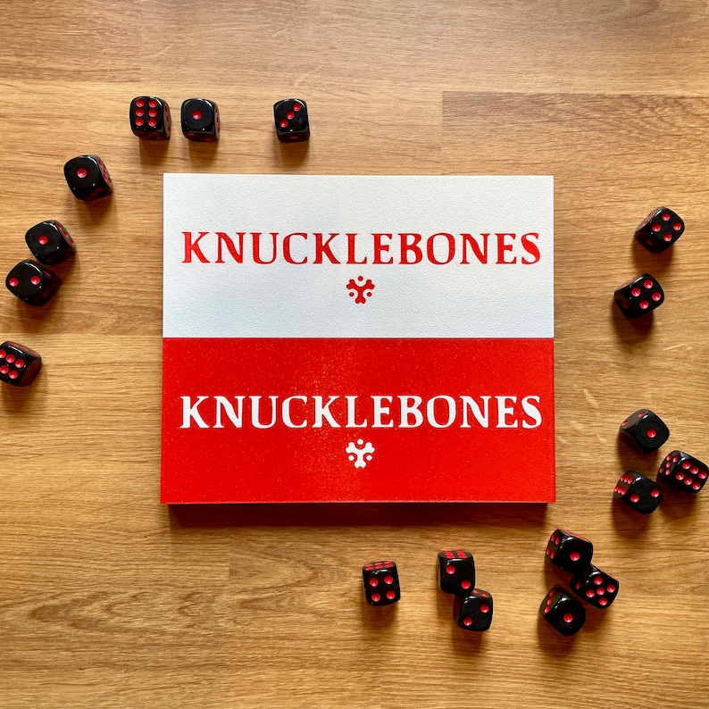 Może przedstawiać: Czerwono-białe pudełko z nadrukiem "KnuckleBones". Pudełko jest otoczone czarnymi kostkami do gry z czerwonymi kropkami.