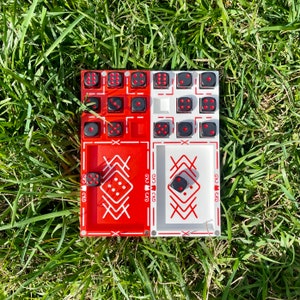May include: A red and white wooden game board with black dice. The board has two sections, each with a different pattern. The dice are arranged in a grid pattern on the board.
