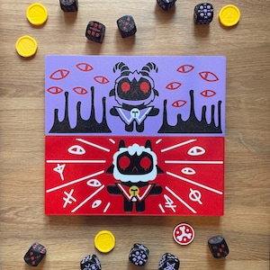 May include: Two rectangular wooden blocks featuring cartoon goat characters. The top block is lavender with a black goat, red eyes, and floating red eyes. The bottom block is red with a white-haired goat, red eyes, white lines, and symbols.