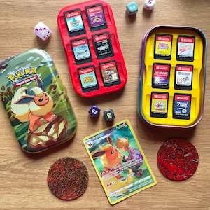 Può includere: Una collezione di accessori e oggetti da collezione per il gaming, tra cui una custodia rossa con cartucce di gioco per Nintendo Switch, una custodia gialla, dadi, una carta collezionabile Pokémon e una moneta. La scatola Pokémon presenta un'illustrazione di un personaggio Eevee.
