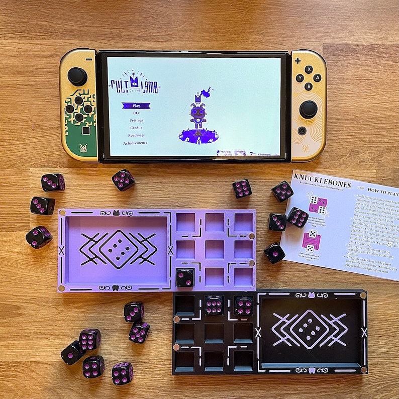 Może przedstawiać: Fioletowo-czarna plansza do gry z kostkami i konsolą Nintendo Switch wyświetlającą grę "Cult of the Lamb". Plansza do gry ma fioletową ramkę i czarne centrum z białym wzorem. Kostki są fioletowe z czarnymi kropkami. Konsola Nintendo Switch jest złota i czarna z białym ekranem.