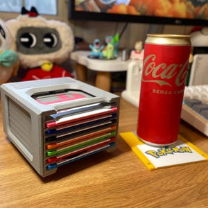 Könnte beinhalten: Ein grauer 3,5-Zoll-Disketten-Organizer mit einem Stapel bunter Disketten. Eine rote Coca-Cola-Dose mit goldenen Akzenten steht auf einem gelb-weißen Pokémon-Untersetzer. Weitere Spielzeuge sind im Hintergrund zu sehen.