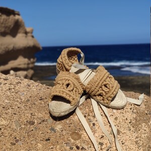 Op de afbeelding: Een paar zandkleurige geweven sandalen met witte veters. De sandalen liggen op een rotsachtig oppervlak met een blauwe zee en lucht op de achtergrond.