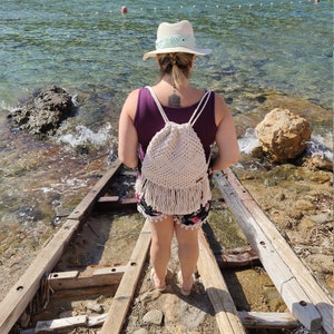 May include: A woman wearing a white hat and a purple tank top stands on a wooden walkway. She is carrying a cream-coloured crocheted drawstring bag with fringe.