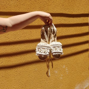 Op de afbeelding: Een paar witte gehaakte sandalen met witte en groene bloemenversieringen. De sandalen hebben een geweven band en een platte zool.