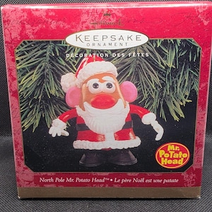 Könnte beinhalten: Ein Weihnachtsschmuckstück mit Mr. Potato Head als Weihnachtsmann. Das Ornament befindet sich in einer roten Schachtel mit der Aufschrift "Keepsake Ornament" und "North Pole Mr. Potato Head". Das Ornament hat einen roten Weihnachtsmannanzug und -hut.