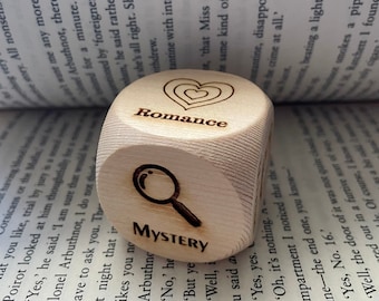 May include: A wooden cube with three sides showing different words: "Romance", "Mystery", and a magnifying glass. The cube is sitting on top of an open book.