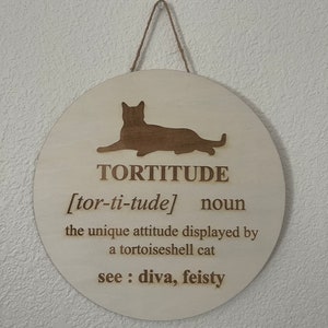 May include: A round wooden sign with a cat silhouette and the word "TORTITUDE" defined as "the unique attitude displayed by a tortoiseshell cat". The definition also includes "see: diva, feisty".