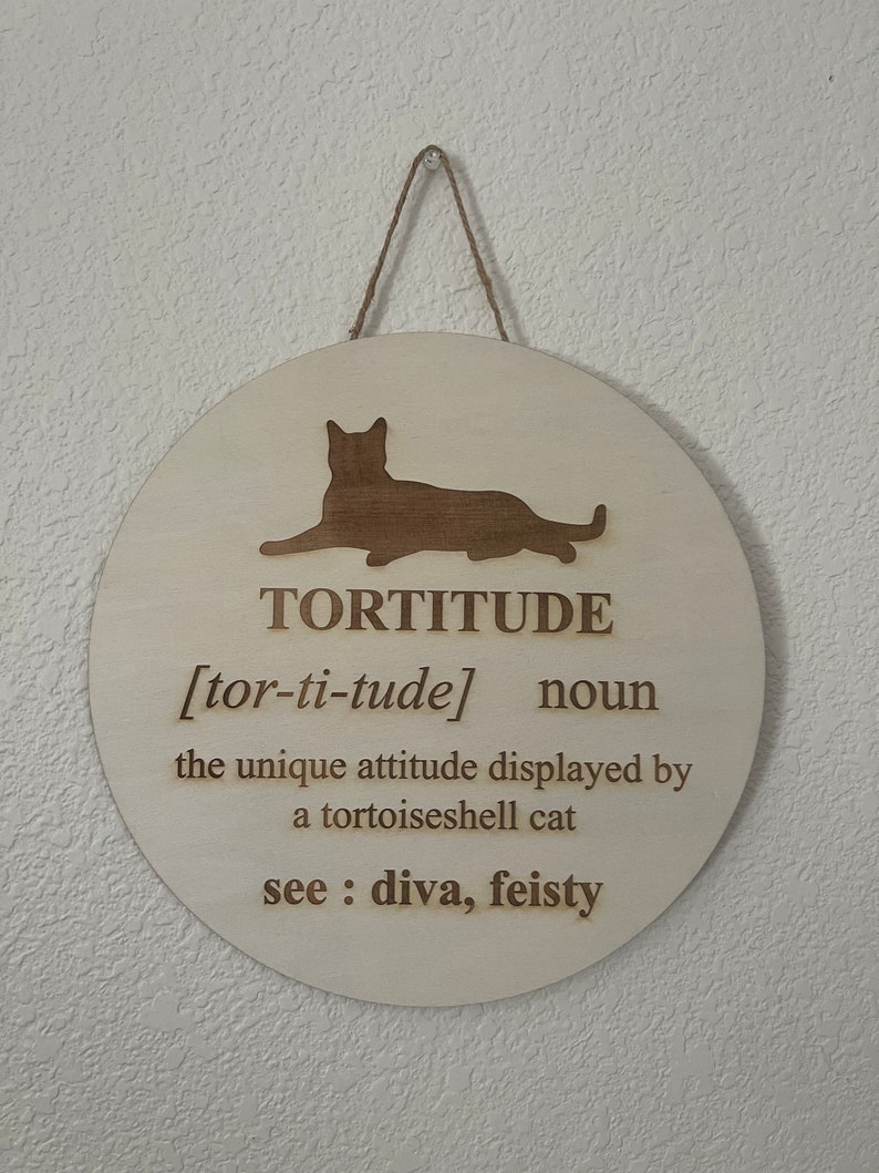 May include: A round wooden sign with a cat silhouette and the word "TORTITUDE" defined as "the unique attitude displayed by a tortoiseshell cat". The definition also includes "see: diva, feisty".