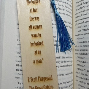 May include: A wooden bookmark with a blue tassel. The bookmark features a quote from F. Scott Fitzgerald's "The Great Gatsby": "He looked at her the way all women want to be looked at by a man."