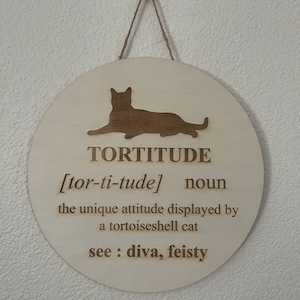 May include: A round wooden sign with a cat silhouette and the word "TORTITUDE" defined as "the unique attitude displayed by a tortoiseshell cat". The sign also says "see: diva, feisty".